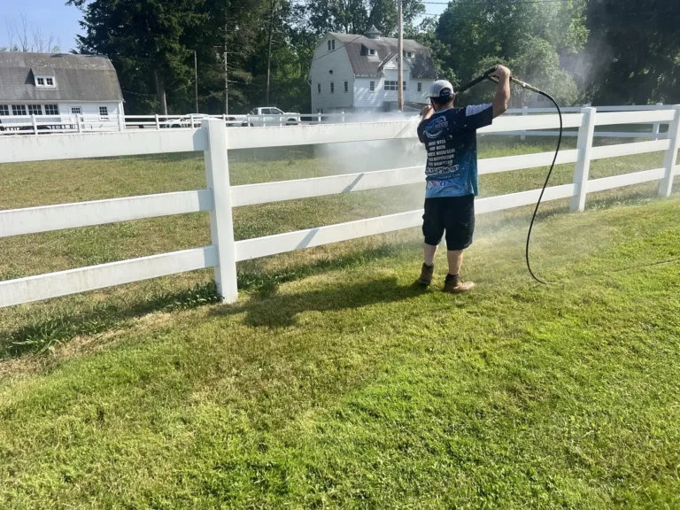 A GrimeBusters Soft Wash and Holiday Lighting Team Member Washes a White Pickett Fence on Bright, Vibrant Grass.